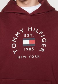Felpa in cotone colore bordeaux con un grande logo circolare bianco che presenta la scritta "TOMMY HILFIGER EST. 1985 NEW YORK" e un piccolo emblema rosso, bianco e blu.