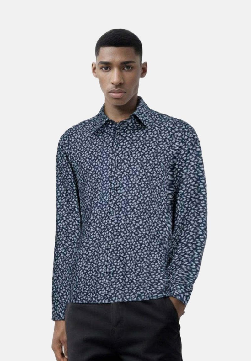 Camicia a maniche lunghe di colore blu navy con piccolo motivo floreale bianco, con colletto e chiusura a bottoni, realizzata in tessuto morbido, indossata con pantaloni scuri.