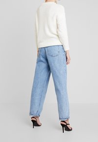 Femme portant un pull en maille blanc, un jean large taille haute bleu clair et des sandales à talons noires, debout devant un fond uni.