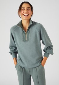 Pullover verde-blu chiaro con colletto alto e zip dorata. Tessuto morbido e liscio con maniche raglan e vestibilità rilassata. Abbinato a pantaloni coordinati.