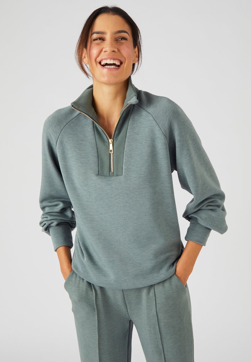 Pullover verde-blu chiaro con colletto alto e zip dorata. Tessuto morbido e liscio con maniche raglan e vestibilità rilassata. Abbinato a pantaloni coordinati.