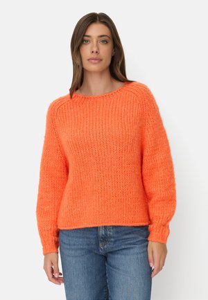 Kvinde iført en lys orange strikket sweater og blå jeans, stående foran en ensfarvet lys baggrund.