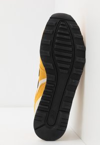 Gul sportskor i mocka med svart texturerad gummisula, vita detaljer och en slank, ergonomisk form.