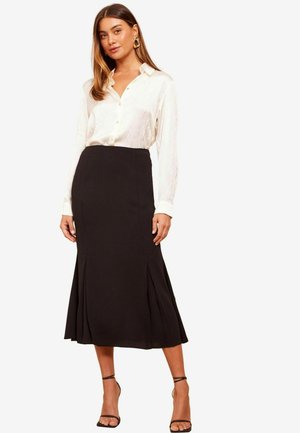 Schwarzer Midirock mit Falten, tailliertem Bund, kombiniert mit einer leichten, langärmeligen weißen Bluse mit Knopfleiste. Riemchenabsätze.