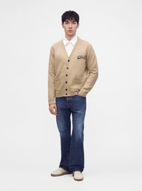Jeune homme portant un cardigan beige boutonné, une chemise blanche, un jean bleu et des baskets de couleur claire, debout avec une main dans la poche.