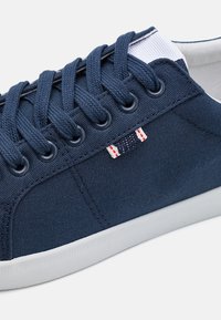 Sötétkék vászon sneaker hat fűzőlyukkal, fehér gumi talppal és egy kis piros-fehér címkével. Texturált szövet és kontrasztos fehér sarok díszítéssel.