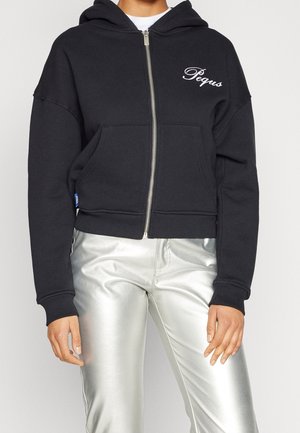 Schwarzer Zip-Hoodie mit weißem „Pegus“-Schriftzug auf der linken Brust, getragen mit glänzenden silbernen Hosen an einer stehenden Person.