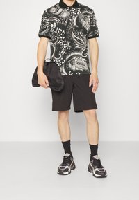 Czarna i biała koszula w wzór paisley z krótkim rękawem, w połączeniu z czarnymi szortami i masywnymi sneakersami, trzymająca czarną torbę.