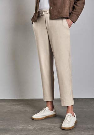 Beige maßgeschneiderte Hose mit Gürtel, kombiniert mit weißen Turnschuhen und einer braunen Jacke auf einem Betonboden vor einer schlichten Wand.