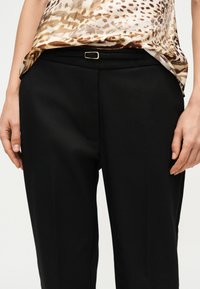 Άτομο που φοράει μαύρο ριχτό παντελόνι με λεπτή μαύρη ζώνη και μπλούζα με λεοπάρ σχέδιο, μπαλωμένη στη μέση.