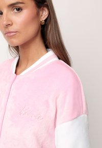 Chaqueta bomber de felpa rosa con mangas blancas, que presenta el logotipo bordado de "Barbie" y un cuello acanalado deportivo. Textura suave y suave brillo.