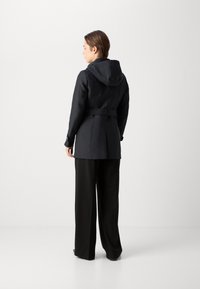 Zwarte trenchcoat met een capuchon, een ceintuur in de taille en knoopmanchetten, gecombineerd met zwarte broek met wijde pijpen. Gladde textuur, strakke uitstraling.