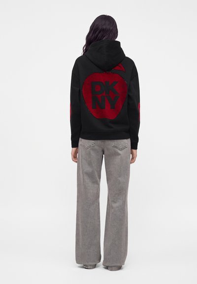 Personne portant un sweat à capuche noir avec une grande pomme rouge et le texte "DKNY" dans le dos, assorti à un jean gris ample.