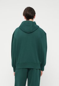 Zelená mikina s velkou kapucí, spadlými rameny a žebrovanými manžetami. Vyrobena z měkké tkaniny, má jednoduchý design bez vzorů.