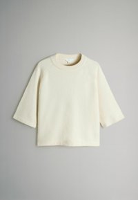 Off-white, pehmeäpintainen neule, jossa on pyöreä kaula-aukko, kolmannesraglan-hihat ja suora helma.