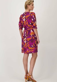 Paarse jurk met abstracte oranje en crèmekleurige print, geaccentueerd op de taille, met gatherings aan de manchetten, gecombineerd met oranje hoge hakken.