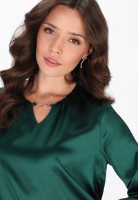 Grüne Satinbluse mit V-Ausschnitt und gerafftem Halsdetail, mit einem subtilen goldenen Kettenakzent am Kragen. Glatte Textur.