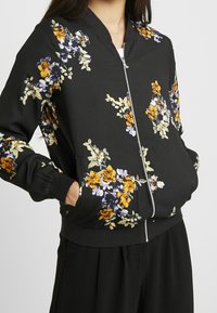 Black bomber jacket featuring floral embroidery in yellow, purple, and white with a zip front, ribbed cuffs, and a relaxed fit.