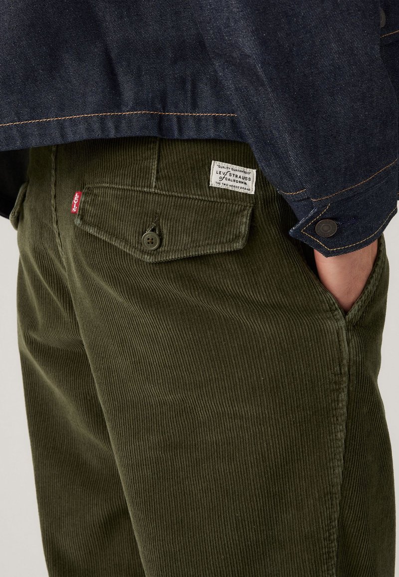 Green corduroy pants with a ribbed texture, featuring two rear pockets and a Levi's label. Paired with a dark denim jacket.