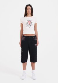 Pantalones cortos de mezclilla negra con un diseño gráfico de calavera, con bolsillos florales coloridos y zapatillas blancas. La camiseta es de color rosa claro con detalles impresos.