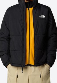 Chaqueta de plumas negra con un cuello alto, con un logo en el pecho, superpuesta a una capa intermedia de forro polar amarillo brillante, combinada con pantalones beige.