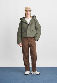 Zaļš, polsterēts jaku ar kapuci, divas priekšējās kabatas un pogu aizdare. Saskaņots ar brūnām biksēm un baltu apavu. Neitrāla fona.