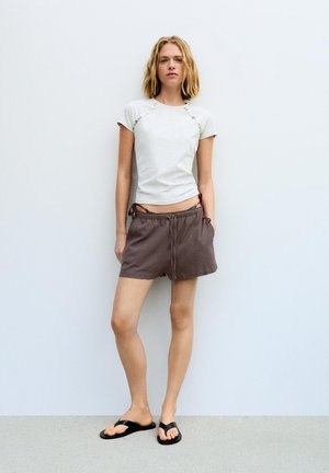 Ung kvinde i hvid top med korte ærmer og brune shorts med hænderne i lommerne, stående mod en ensfarvet lys væg iført sorte flip-flops.