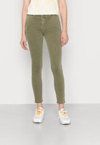 Jeans ajustados de color verde oliva con una textura suave, diseño de cinco bolsillos y cierre con cremallera. Combinados con zapatillas blancas que tienen cordones naranjas.