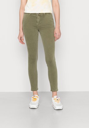 Jeans ajustados de color verde oliva con una textura suave, diseño de cinco bolsillos y cierre con cremallera. Combinados con zapatillas blancas que tienen cordones naranjas.
