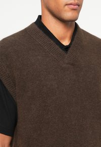 Άνδρας που φοράει ένα σκούρο καφέ φούσκα V-λαιμό γιλέκο πάνω από ένα μαύρο πουκάμισο με κολάρο, δείχνοντας το λαιμό και μερικό πρόσωπο.