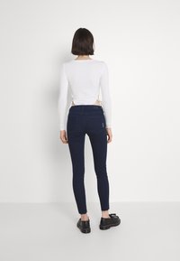 Top bianco a maniche lunghe con apertura sulla schiena abbinato a jeans skinny blu scuro. La modella indossa scarpe nere; tessuto liscio e design aderente.