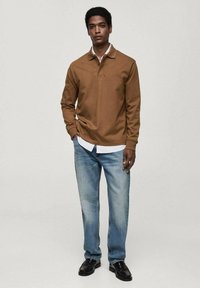 Hnědá dlouhá polo košile, směs bavlny, s dvouknoflíkovým zapínáním, přeložená přes bílou košili s límečkem, kombinovaná se světlými modrými džínami a černými botami.