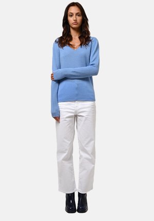 Helle blaue, langärmelige Pullover aus weichem Material mit V-Ausschnitt. Kombiniert mit weißen, weit geschnittenen Hosen und schwarzen Ankle Boots.