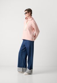 Una persona indossa occhiali da sole neri, una felpa leggera di colore rosa pallido, jeans ampi blu con i risvolti e sneakers bianche, in piedi davanti a uno sfondo chiaro uniforme.