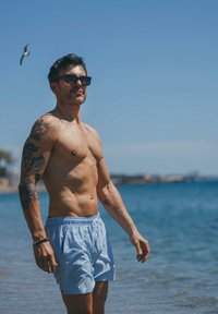 Mężczyzna w niebiesko-białych pasiastych kąpielówkach, w okularach przeciwsłonecznych, z tatuażami, stojący na plaży z jasnym niebem i wodą.