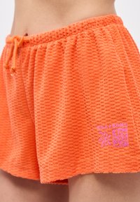 Πορτοκαλί υφασμάτινες βερμούδες terry με ελαστική μέση και κορδόνι, με ροζ λογότυπο Billabong και σχέδια λουλουδιών στο μπροστινό μέρος.