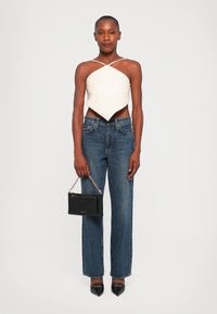 Witte, asymmetrische crop top met dunne bandjes, gecombineerd met rechte blauwe jeans. Houdt een kleine, zwarte handtas met een kettingriem vast. Zwarte hakken.