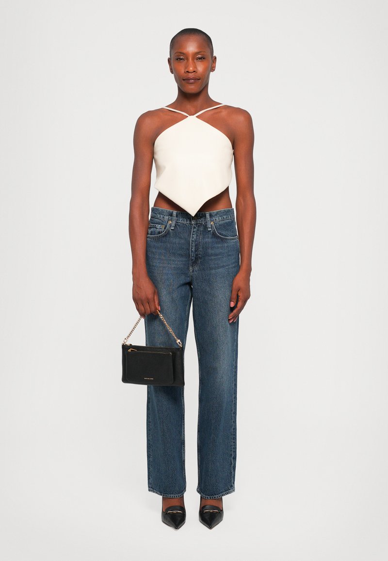Witte, asymmetrische crop top met dunne bandjes, gecombineerd met rechte blauwe jeans. Houdt een kleine, zwarte handtas met een kettingriem vast. Zwarte hakken.