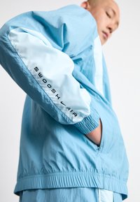 Ljusblå jacka med elastiska ärmslut och avslappnad passform. Har "SWOOSH AIR" tryckt på ärmen, tillverkad av ett slätt, lättviktsmaterial.