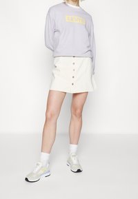 Fioletowa bluza z żółtym logo "LEVI'S", biała spódnica jeansowa na guziki, oraz beżowe sneakersy z pastelowymi akcentami i białe skarpetki.