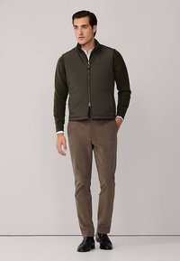 Herrengeflettete Weste in Dunkelgrün, darüber ein langärmliges Hemd getragen. Braune Hose und schwarze Schuhe vervollständigen das Outfit. Glatte Textur, figurbetontes Design.