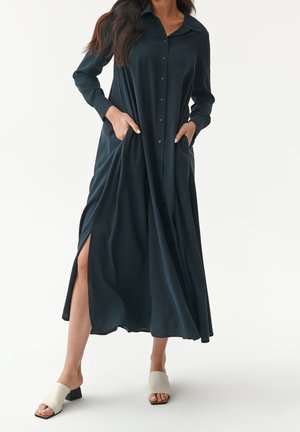 Vestido camisa largo de color azul marino oscuro con botones en la parte delantera, cuello, bolsillos laterales y altas aberturas. Combinado con sandalias de tacón cuadrado de color claro.