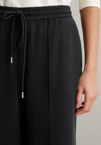 Gros plan sur une personne portant un pantalon noir à cordon et un haut à manches longues crème, avec une main détendue à ses côtés.