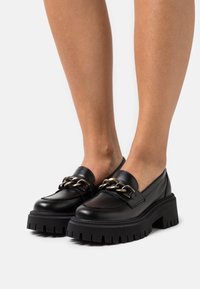 Mocassins en cuir noir avec une semelle épaisse et côtelée, ornés d'une chaîne dorée décorative sur le dessus pour un détail supplémentaire.