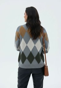 Kobieta o długich ciemnych włosach ma na sobie szary sweter w romby w kolorach brązu, bieli i ciemnej zieleni, czarne spodnie oraz brązową torbę na ramię.