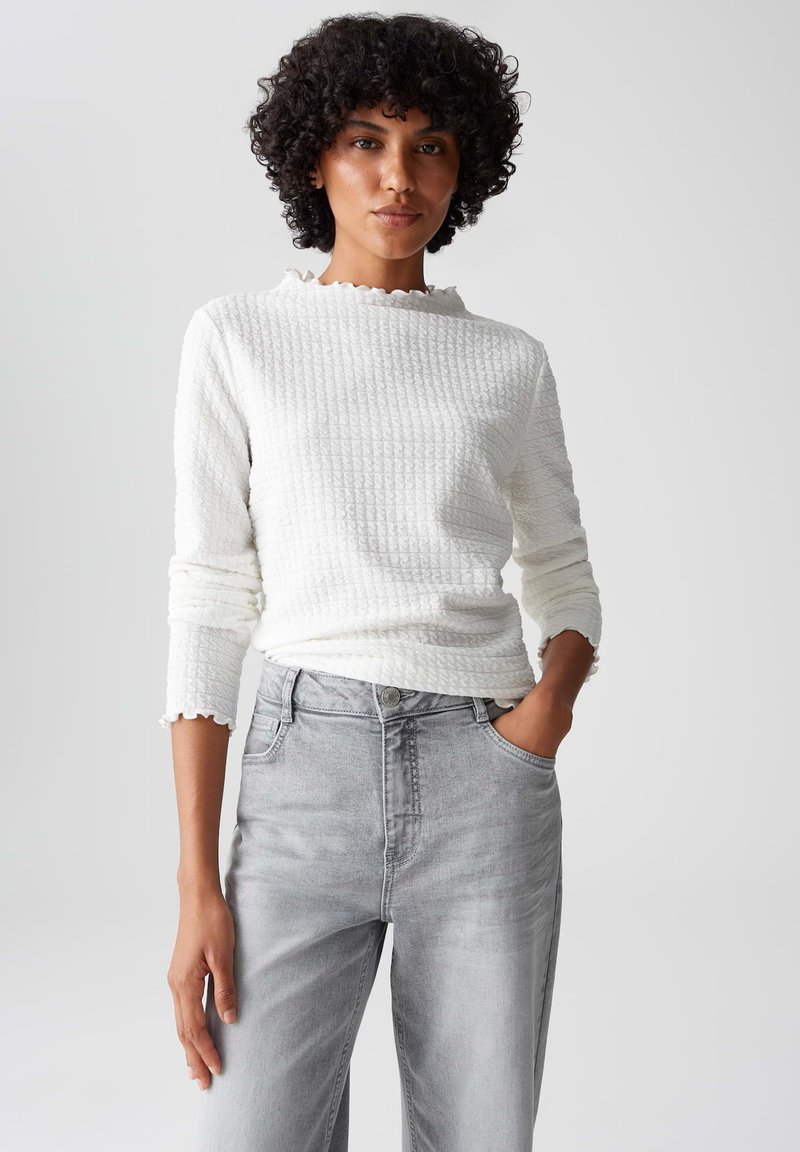Frau mit lockigem Haar, die ein strukturiertes weißes Langarmoberteil und eine hellgraue High-Waist-Jeans trägt, Hand in der Tasche, neutraler Hintergrund.
