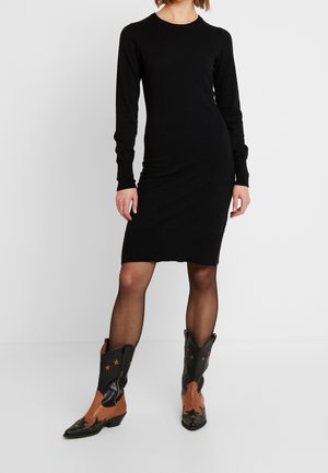 Femme portant une robe noire ajustée à manches longues, associée à des bottines western noires et marron avec des motifs d’étoiles.