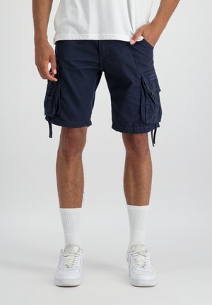 Man wearing navy blue cargo shorts, white crew socks, and white sneakers, standing with one hand in pocket against plain background.