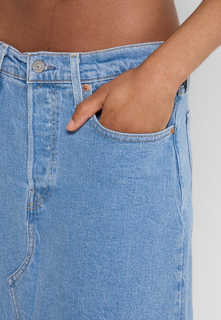 Mano colocada dentro del bolsillo derecho de unos jeans de mezclilla azul claro, mostrando la cintura y parte del abdomen contra un fondo liso.