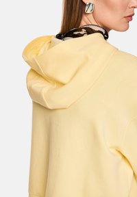 Gelber Hoodie aus weichem Material, mit großzügiger Kapuze, entspanntem Schnitt und einem mehrlagigen Kragen mit schwarzem und beigem Muster.